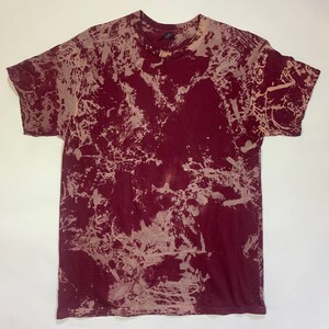 May include: A burgundy t-shirt with a unique bleach-dye pattern. The short-sleeved shirt features a crew neck and a design of light beige splotches across the fabric. The overall effect is a marbled, abstract design, perfect for casual wear.