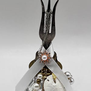 May include: A decorative bottle stopper featuring a repurposed fork, adorned with a white ribbon, pearl accents, a gold tassel, and a white sand dollar. The stopper is designed to fit a glass bottle.
