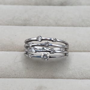May include: A silver ring with multiple bands, each adorned with clear, round gemstones of varying sizes. The ring has a modern, minimalist design, with the gemstones set along the bands.