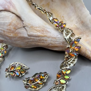 May include: A gold-tone necklace and matching clip-on earrings set featuring a leaf motif. Each leaf is accented with marquise-cut iridescent orange and yellow rhinestones. The metalwork has a textured finish, and the necklace includes a chain.