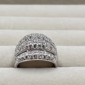 May include: A silver-toned ring with a domed top, encrusted with numerous small, clear gemstones. The ring has a wide band and a layered design, creating a sparkling, eye-catching effect. The gemstones are tightly set, covering the entire surface of the ring.