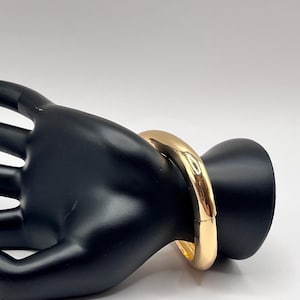 May include: A gold-colored bangle bracelet. The bracelet is a thick, rounded design with a smooth, polished surface. The bracelet is shown on a black hand-shaped display, set against a white background.