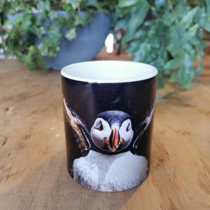 May include: A black ceramic mug with a colorful image of a puffin with its wings spread out. The puffin is looking directly at the camera.