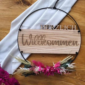 May include: A wooden sign with the text "Herzlich Willkommen" in a black metal hoop with dried flowers.