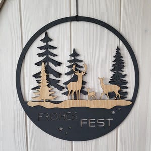 Peut inclure: Un cercle en bois noir avec un motif découpé représentant une scène de forêt d'hiver avec des cerfs et le texte "Frohes Fest".