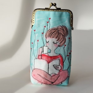 May include: A light blue fabric coin purse with a brass clasp. The purse features an embroidered design of a woman sitting with a book and a cup of coffee. The background is a light blue with red hearts.