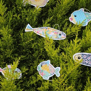 May include: A collection of colorful, fabric fish ornaments with various patterns and designs, including polka dots and floral prints. The fish are suspended against a backdrop of green foliage, creating a whimsical and decorative display.