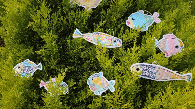 May include: A collection of colorful, fabric fish ornaments with various patterns and designs, including polka dots and floral prints. The fish are suspended against a backdrop of green foliage, creating a whimsical and decorative display.