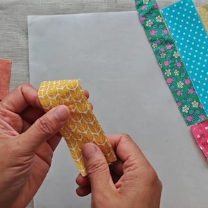 May include: A person holding a yellow fabric strip with a leaf pattern. Various fabric strips with floral, polka dot, and other patterns are arranged on a white surface, suggesting a quilting or sewing project.