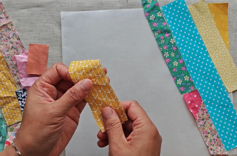 May include: A person holding a yellow fabric strip with a leaf pattern. Various fabric strips with floral, polka dot, and other patterns are arranged on a white surface, suggesting a quilting or sewing project.