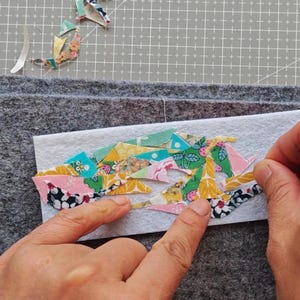 May include: A craft project in progress, featuring a white felt rectangle with colorful fabric scraps being arranged. The scraps have floral and geometric patterns in pink, yellow, and blue. Gray felt squares are in the background.