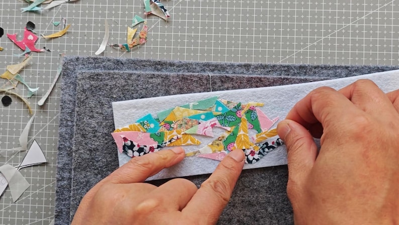 May include: A craft project in progress, featuring a white felt rectangle with colorful fabric scraps being arranged. The scraps have floral and geometric patterns in pink, yellow, and blue. Gray felt squares are in the background.