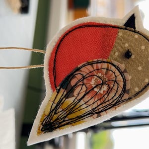 May include: A handmade bird-shaped fabric ornament. The bird features a red and tan polka-dot body, with a floral patterned wing and a black stitched outline. The bird is attached to a twine string with two green beads.
