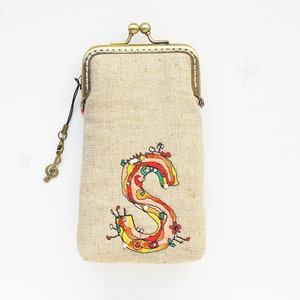 May include: A small beige fabric pouch with a brass clasp and a music note charm. The pouch has an embroidered colorful letter "S" on the front.