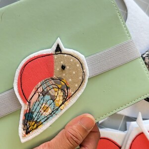 May include: A handmade bird-shaped patch with red, tan, and floral fabric details is affixed to a light green notebook. The patch is outlined in black stitching. A gray elastic band secures the notebook. Scissors with a black and white checkered handle are in the background.