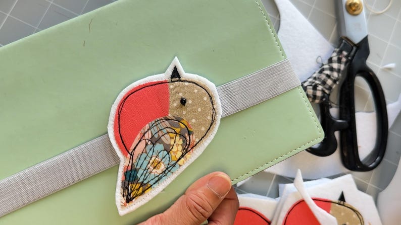 May include: A handmade bird-shaped patch with red, tan, and floral fabric details is affixed to a light green notebook. The patch is outlined in black stitching. A gray elastic band secures the notebook. Scissors with a black and white checkered handle are in the background.