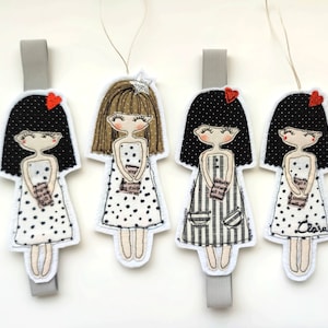 May include: Four handmade fabric doll bookmarks with black hair, brown hair, and black and white polka dot dresses. Each doll is holding a book with text on the cover. The bookmarks are attached to gray ribbon.