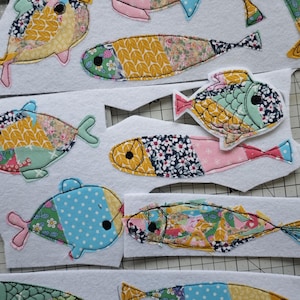May include: A collection of colorful, fabric fish appliqu&eacute;s in various sizes and patterns. The fish feature floral, polka dot, and geometric designs in shades of blue, pink, yellow, and green. Each fish is outlined with black stitching.