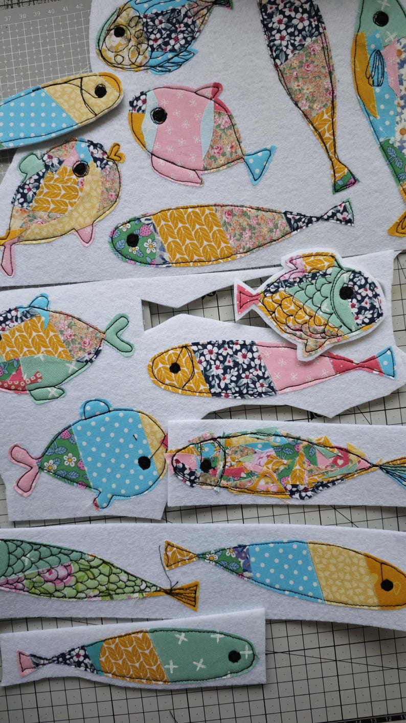 May include: A collection of colorful, fabric fish appliqu&eacute;s in various sizes and patterns. The fish feature floral, polka dot, and geometric designs in shades of blue, pink, yellow, and green. Each fish is outlined with black stitching.
