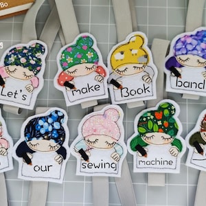 May include: Eight colorful fabric-covered bookmarks with different designs. Each bookmark features a cartoon character with a pencil and a message. The messages are: Let's, make, book, band, with, our, sewing, machine.