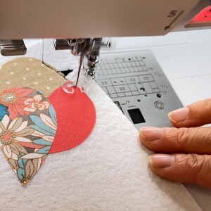 May include: A sewing machine stitching a heart-shaped applique onto white fabric. The applique is composed of three sections: a red circle, a floral pattern, and a tan polka dot design. The machine's needle is in action, guided by hands.
