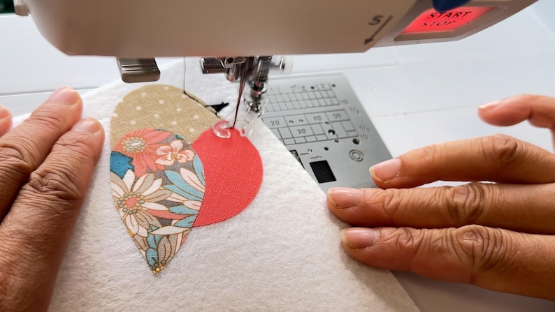 May include: A sewing machine stitching a heart-shaped applique onto white fabric. The applique is composed of three sections: a red circle, a floral pattern, and a tan polka dot design. The machine's needle is in action, guided by hands.