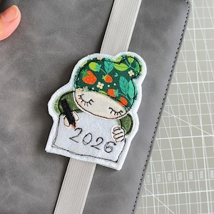 May include: A gray notebook with a light gray elastic band and a decorative felt bookmark. The bookmark features a cartoon gnome holding a pen and a sign with the year 2026. The gnome wears a green hat with orange and white accents.