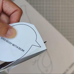 May include: Scissors cutting a white paper bird body template with the text "BIRD BODY WITH BEAK". The template is on a white surface, with a grid pattern visible in the background.