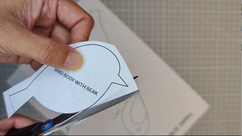 May include: Scissors cutting a white paper bird body template with the text "BIRD BODY WITH BEAK". The template is on a white surface, with a grid pattern visible in the background.
