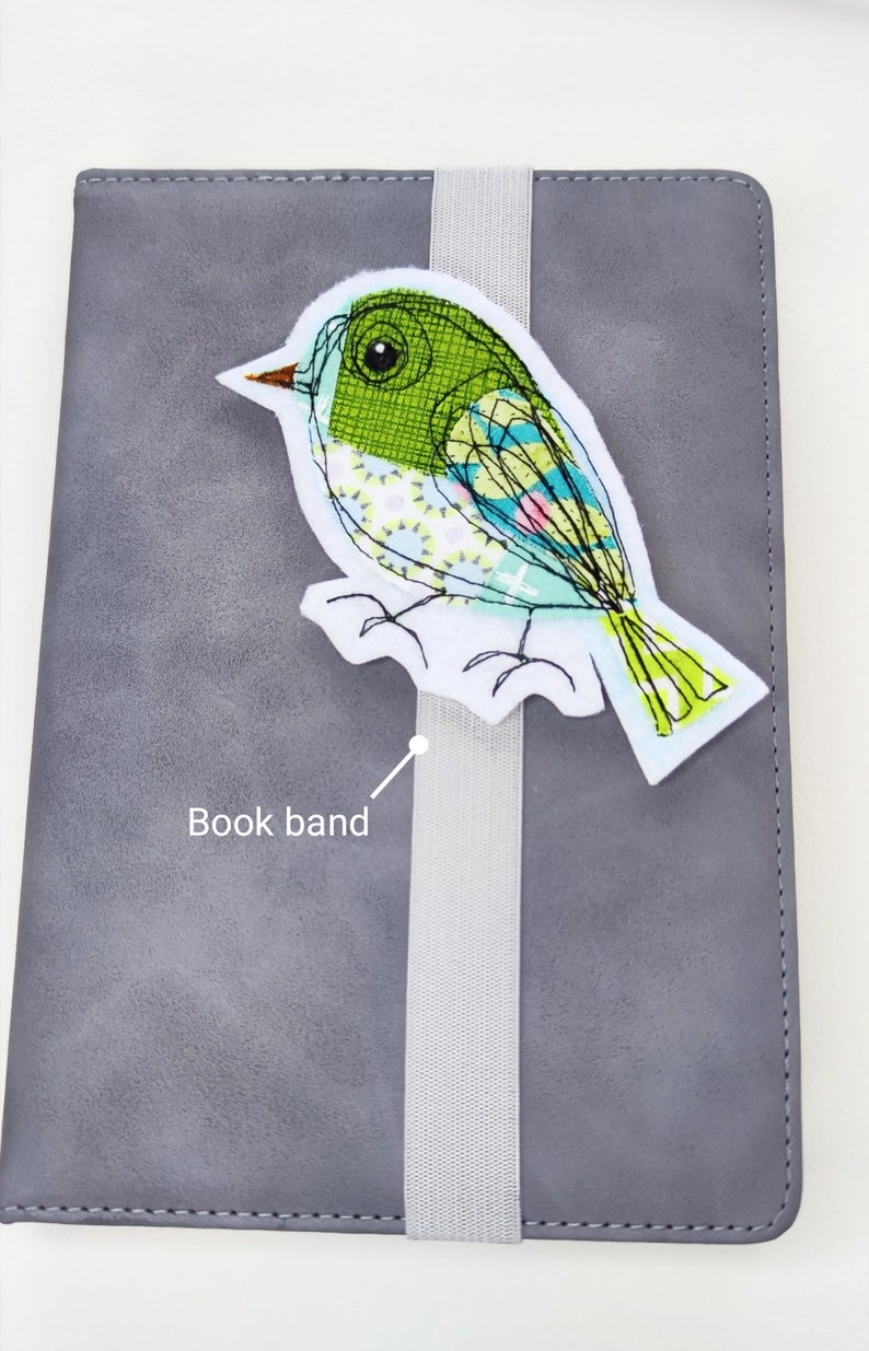 May include: A gray notebook with a decorative bird bookmark. The bird is white with green, blue, and yellow accents. A gray elastic book band holds the notebook closed.