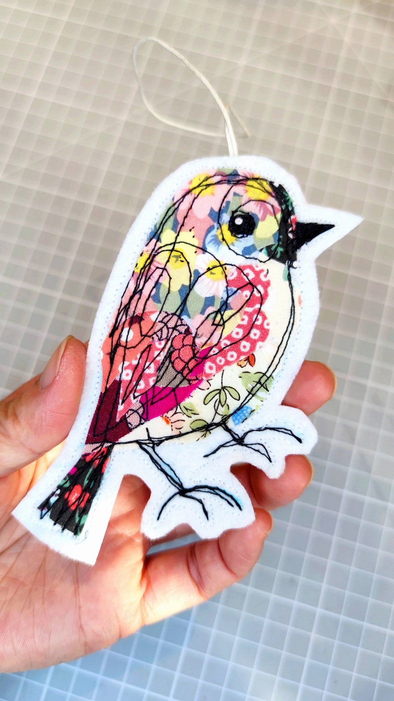 May include: A colorful fabric bird ornament with a white felt backing and black stitched details. The bird features a patchwork design with floral patterns in pink, yellow, and red. A white string is attached for hanging.