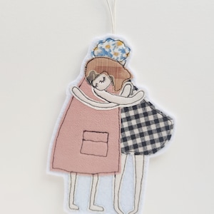 Puede incluir: Dos figuras de fieltro de chicas abrazándose. La chica de la izquierda lleva un vestido rosa con un bolsillo y la chica de la derecha lleva un vestido a cuadros blanco y negro. Ambas chicas tienen el pelo castaño y los ojos cerrados.