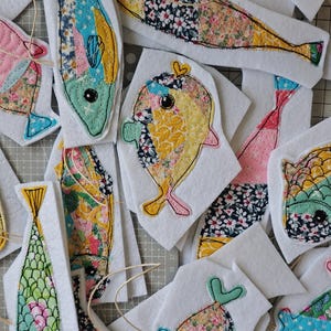 May include: A collection of colorful, fabric fish appliqu&eacute;s with various patterns and colors, including floral and geometric designs. Each fish is outlined with black stitching and attached to a white felt backing. Some have string attached for hanging.