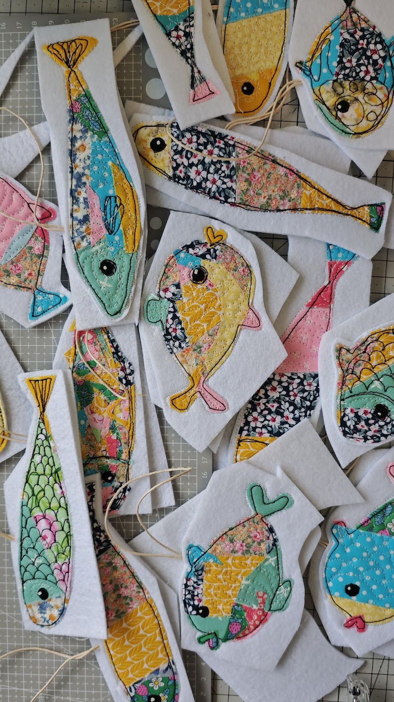 May include: A collection of colorful, fabric fish appliqu&eacute;s with various patterns and colors, including floral and geometric designs. Each fish is outlined with black stitching and attached to a white felt backing. Some have string attached for hanging.