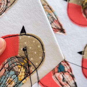 May include: Close-up of a hand-stitched bird applique on white fabric. The bird features red, tan, and patterned fabric details, with black stitching outlining the design. The needle and thread are visible, indicating the craft in progress.