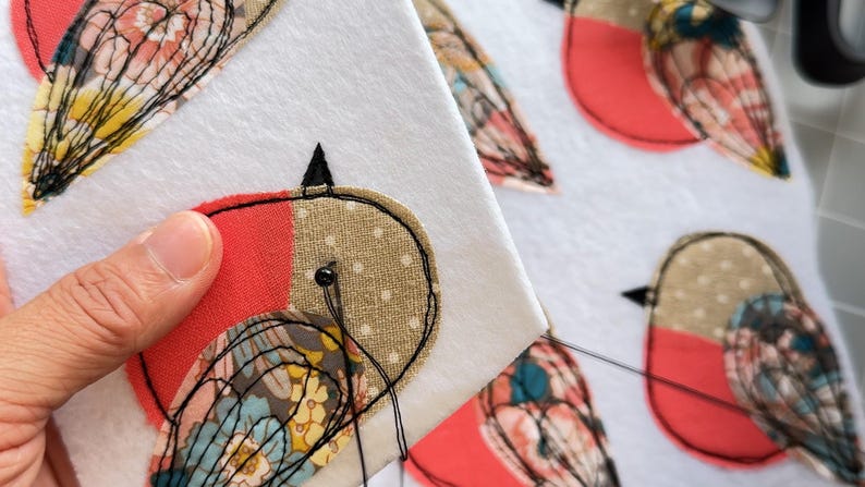 May include: Close-up of a hand-stitched bird applique on white fabric. The bird features red, tan, and patterned fabric details, with black stitching outlining the design. The needle and thread are visible, indicating the craft in progress.