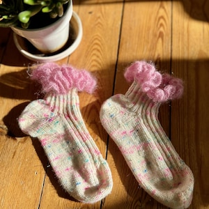 May include: A pair of hand-knitted socks with a speckled pattern in white, pink, and blue. The cuffs are a fluffy pink, and the socks are displayed on a wooden surface. The socks are designed for comfort and warmth.