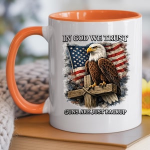 May include: White ceramic mug with an orange handle and interior. The mug features an American flag and a bald eagle perched on a wooden cross. Text on the mug reads "IN GOD WE TRUST" and "GUNS ARE JUST BACKUP."