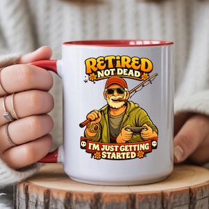 May include: White ceramic mug with a red interior and handle. The mug features a cartoon illustration of a retired fisherman with the text "Retired Not Dead" and "I'm Just Getting Started". The mug is held by a person.