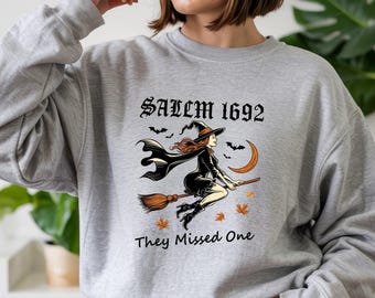 May include: A light gray crewneck sweatshirt featuring a graphic of a witch flying on a broomstick. The text reads SALEM 1692 and They Missed One. The design includes a crescent moon, bats, and orange autumn leaves in a black, orange, and white color palette.