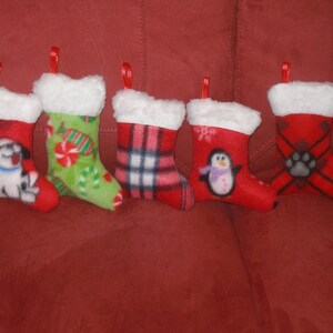 May include: Five red Christmas stockings with white fur trim. Each stocking features a different design: a dog, candy canes, plaid, a penguin, and a paw print.