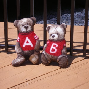 May include: Two plush teddy bears wearing red sweaters with white letters. The bear on the left has the letter "A" on its sweater, and the bear on the right has the letter "B" on its sweater.