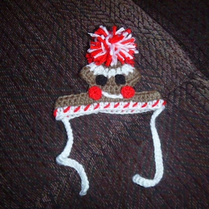 May include: A brown crocheted gingerbread man hat with a white pom-pom and red and white candy cane trim. The hat has a white crocheted strap.