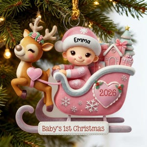 May include: A Christmas ornament featuring a reindeer with a floral crown pulling a pink sleigh. Inside the sleigh sits a baby wearing a pink hat with the name Emma. The sleigh displays a heart with 2026 and a sign reading Baby's 1st Christmas.