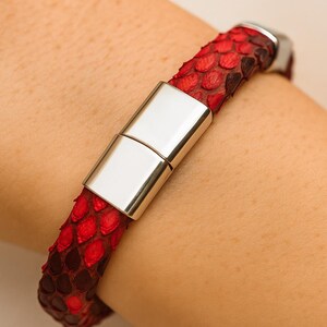 May include: Red and black snakeskin bracelet with a silver magnetic clasp. The bracelet is wrapped around a wrist, showcasing the texture and color variations of the snakeskin. The clasp is rectangular and shiny.