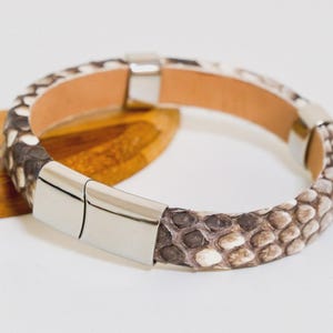 Könnte beinhalten: Ein Schlangenlederarmband mit Magnetverschluss. Das Armband weist ein Muster aus braunen, grauen und weißen Schuppen auf. Die Innenseite ist hellbraunes Leder. Der Verschluss ist silberfarben glänzend.
