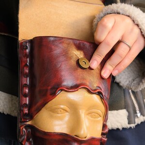 Peut inclure: Un sac en cuir artisanal au design unique. Le sac présente un visage sculpté sur le devant, avec une palette de couleurs bordeaux et beige. Il possède un fermoir magnétique et une bordure cousue. Le sac est en cuir de haute qualité.