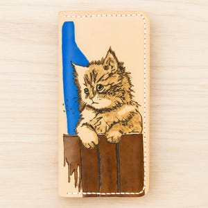 May include: A beige leather phone case featuring a hand-painted illustration of a fluffy brown and tan kitten. The kitten is sitting in a brown box with a blue shape on the left side. The case has white stitching around the edges.