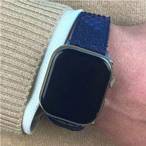 May include: A silver smartwatch with a dark blue screen and a textured blue strap. The watch is worn on a wrist, with a light blue shirt cuff and a beige sweater sleeve in the background.