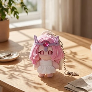 May include: A plush doll with pink hair, a purple crown, and a white dress. The doll has a keychain attached. The doll is on a wooden table.